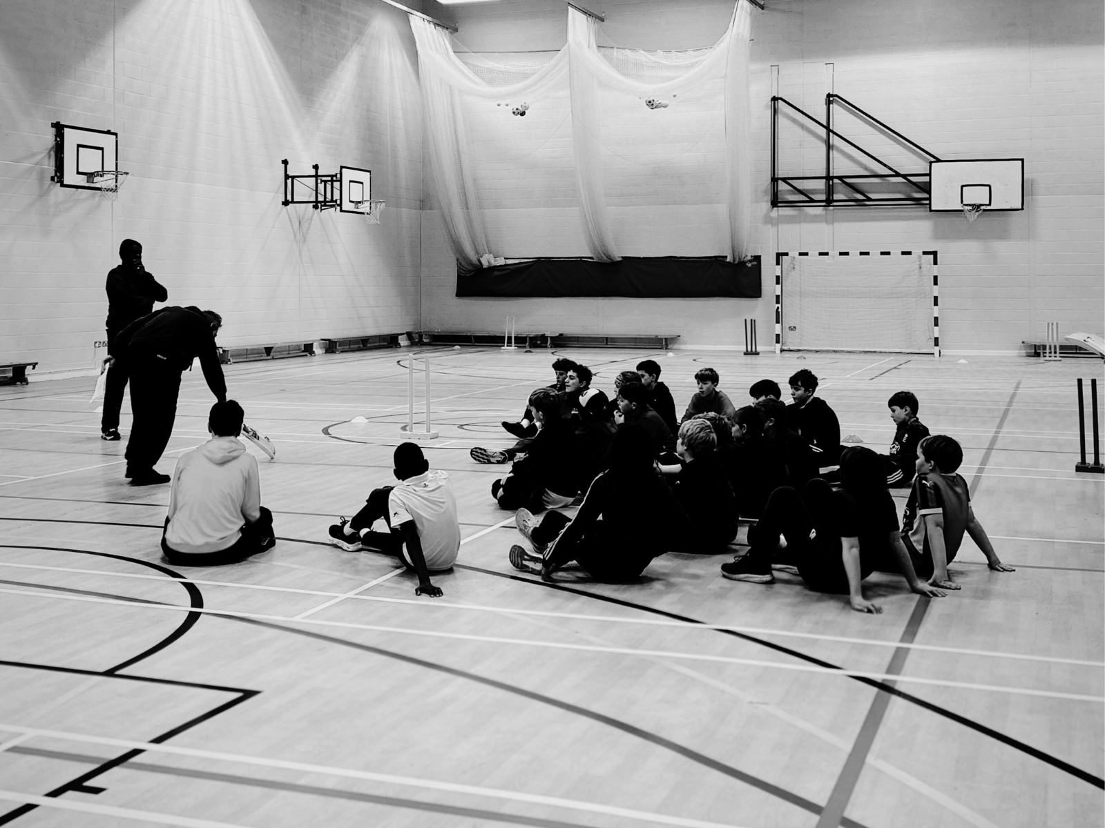 Young cricket players seated on a school sports hall floor listening to coaches, with cricket stumps set up in a multi-purpose gymnasium